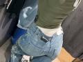MargoFun's ass in tight blue jeans with a black belt, green top, white underwear visible, standing in a store with a denim jacket on a hanger.