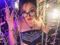 LunaixMoon with glasses, red lipstick, black bandage top, holding chains, looking up, showing tits, tattoo on chest, dark hair, purple and orange lighting, and a confident expression.