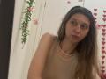 Sanella has long brown hair, a pearl necklace, and a beige sleeveless top, with a neutral expression, in a slightly angled selfie, showing her tits and ass.