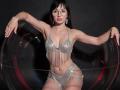 Rouzi mit Brille, die ein glitzerndes Bikini-Set trägt, hält eine klare Kugel in der Hand, zeigt ihre Brüste und ihren Hintern, schwarzes Haar, trägt hängende Ohrringe, dunkler Hintergrund, selbstbewusste Pose..
