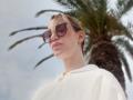 MissFrenchy with blonde hair, wearing large brown sunglasses, white hoodie, and gold earrings, standing in front of a palm tree, looking down with a slight smirk.