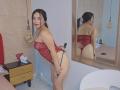 MelaniCoox in a red lace bodysuit, holding a black strap, bending over, showing her ass, with black hair in a ponytail, reflected in a mirror, in a bedroom.