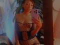 HenutmireRossi takes a selfie in a mirror wearing a black fishnet top that reveals her tits, with her phone in hand, in a dimly lit room.