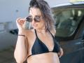 Milagres in a black bikini top, adjusting round sunglasses, with wet, wavy brown hair, wearing a red bracelet and necklace, standing by a black car.