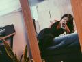 LilyConorr, wearing a black halter dress, lies on a couch, taking a mirror selfie with an iPhone. Her ass is visible, and she has long, wavy brown hair.