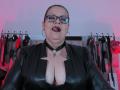 VanesaMoka with glasses, black leather jacket, and choker, tongue out, showing large boobs in a black lace top, hoop earrings, and a pentagram necklace.