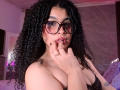 VanessaBlake with curly black hair, glasses, and a black lace bra, biting her finger with her hand on her chest, showing her tits and tattoos on her fingers.
