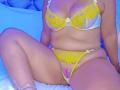AuroraBenson is sitting on a bed wearing yellow lace lingerie. She has big boobs, a chubby belly, and a visible pussy through her sheer yellow panties.