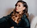 Niferttiti with wavy brown hair, wearing a green satin shirt, black top, and sitting on a white chair, looking to the side with a pouty expression.