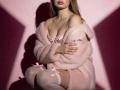 Destinis with pink fluffy jacket, off-shoulder, showing tits, white thigh-highs with pink bows, crouching, hands on jacket, shadowed pink wall.