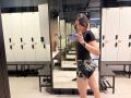 HeikeCarchi with medium tits, black t-shirt, and black/white camo shorts, taking a mirror selfie in a gym locker room, holding a blue phone, wearing a black wristband.