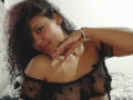 AriaVega with curly black hair, smiling, wearing a sheer black top with polka dots, exposing her small tits, and a gold cross necklace, resting her chin on her hand.
