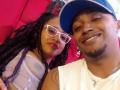 LilithAndAsmodeo with black braids, white glasses, and visible tits, lying next to a smiling black dude with a blue cap, white shirt, and necklace.