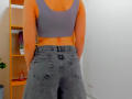Steisii's back, wearing a purple sports bra and gray, slightly baggy jeans, hands in pockets, revealing a small, red, slightly visible butt, in a well-lit room.