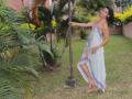 NastyNymphoxxx in a floral halter dress, standing with one arm on a pole, showing her ass and tits, in a grassy yard with palm trees.