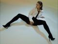 MilagrosEdmisten, a woman with wet, slicked-back hair, sits on a white floor with legs spread wide. She wears a white shirt, black tie, and black pants with high heels. Her expression is seductive.