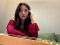 ZulaGullick with long dark hair, red off-shoulder top, looking slightly to the side, hand in hair, sitting at a wooden table with a chessboard in front.