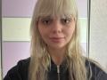DominiqueBly has blonde hair, bangs, and a slight smile. She's wearing a black zip-up, and her pink lips are slightly parted. Her tits are small, and she's cute.