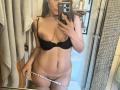 SaraWestt with large, natural tits in a black bra, pulling down white panties, showing her pubic hair, taking a mirror selfie in a bathroom. Her phone covers part of her face.