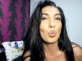 BrunetteBabe69 with long black hair, brown eyes, and pouty lips, making a kissing face, wearing a strapless top, with a pillow featuring blue birds in the background.