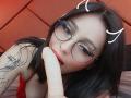 PaisleyMour with glasses, black hair, and tattoos, sucking on a large, pale, circumcised dildo, red lipstick, and a playful, horned emoji overlay.