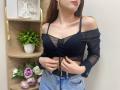 Rioria with bare shoulders, black sheer top, small boobs, tied front, denim jeans, hands on waist, brown hair, gold earrings, subtle pink lipstick, white wall, wooden shelf.
