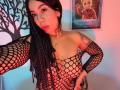 MarianaJackson with black fishnet top, showing her tits through the net, long black braids, red lipstick, tattoo on right arm, necklace, and a "Frozen" poster in the background.
