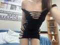 Ramilla in a black, ripped dress, exposing her tits and ass through cut-out sides, standing in a room with blue pillows and a wooden shelf.