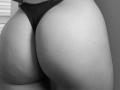 SugarrX's butt in black thong, showing a tight, smooth, round, and slightly scarred ass. The thong is positioned high, highlighting her well-defined, curvy butt. Black-and-white photo.