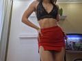 MilaYanis with slim, toned abs, wearing a black lace bralette and a red skirt with a side slit, hands on hips, in a room with a fish tank.