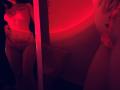 Tisia stands in red lighting, wearing a lace bra and matching panties, with her hand on her hip and a reflection showing her bare ass. Red light casts a sexy glow.