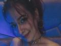 LolyYunno with red hair, hoop earrings, nose ring, and a choker, smiling under blue lighting, showing a slightly open mouth and a small lip piercing.