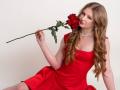 AkilahGhil with long, wavy blonde hair in a red dress, holding a red rose to her nose, wearing a diamond necklace, sitting with her legs slightly apart.