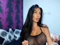 BrunetteBabe69 with long black hair, pouty lips, wearing a black fishnet top that shows her big, round tits. She's holding her finger up, looking at the camera.
