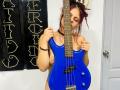 Sophie Hill, con capelli rossi e rosa, che tiene una chitarra elettrica blu, nuda con i seni visibili, unghie rosa, una collana nera e un braccialetto nero, in una stanza molto luminosa..