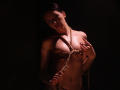 TriciaBarnes with dark hair, topless, and bound with rope, covering her small tits with her hands, a belly button piercing visible, dimly lit, and looking seductively at the camera.