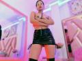 MelisaDaltonn with crossed arms, topless, black star pasties on her tits, wearing a black vinyl skirt, black thigh-highs, standing in a pink and blue lit room.