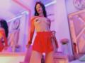 MelisaDaltonn with black hair, red headband, red fishnet sleeves, red skirt, and red pasties covering her tits, standing in a neon-lit room, looking down with a slight smile.