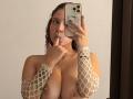 ValeryScot with curly red hair, holding phone, shushing finger near mouth. Wearing white fishnet sleeves, exposing large bare tits. Reflecting in mirror. Red nail polish.