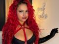 Estefy69 with bright red curly hair, black leather gloves, red harness, and black bra, showing off her big tits and confident smile.