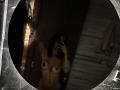 Nude MajoJones with small tits, dark nipples, and a belly button piercing, taking a mirror selfie with arms raised, holding a phone, in a dimly lit bathroom.