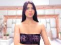 HellenBells tiene el cabello castaño oscuro, lleva un top negro de encaje sin tirantes que deja ver sus pequeños senos y un collar dorado, y está de pie con una ligera sonrisa..