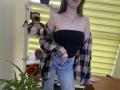 Nesie with bare tits in a black strapless top, pink plaid shirt off her shoulders, and high-waisted jeans, standing in a room with a tiger stuffed animal.