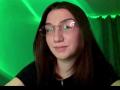SunnyKlusman with long brown hair, wearing clear glasses, black shirt, green-lit background, slight smile, nose piercing, and fair skin.