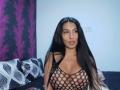 BrunetteBabe69 with long black hair, pouty lips, wearing a black fishnet top that exposes her big tits and nipples, sitting on a bed with geometric pillows.