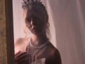 RosaVance with pouty lips, wearing a black lace headpiece, pearl necklace, and off-shoulder white dress, holding her tits, standing in a softly lit, warm-toned room.