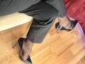 Hotsexydolly's shiny black heels on a wooden floor, black pants, and a red textured cushion in the background. Her legs are crossed, showing off her pointed-toe shoes.
