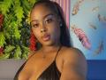 NaomiLoveHot has dark brown skin, full lips, and black hair in a ponytail. She wears a black halter top, showing her small tits, and a gold necklace.