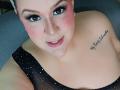 GiiannaMorrys with pink blush, black eyeliner, and white polka dot dress, showing cleavage, tattoo on right shoulder, smiling, and looking at camera. Her tits are partially visible.
