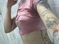 EvaaFarix in a pink shirt, wet and clinging to her tits, showing tattoos on her arm and side, with water droplets on her skin.